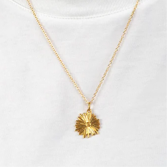 A gold sunburst pendant necklace worn on a white crewneck t-shirt. The pendant has a textured, faceted starburst design and is suspended from a delicate gold chain.