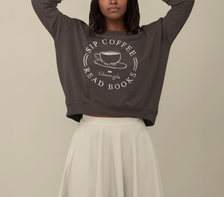 A woman wearing a dark brown cropped sweatshirt with a white graphic print that reads 'SIP COFFEE READ BOOKS' and 'Calmer Girls,' paired with a cream pleated skirt, against a plain beige background.