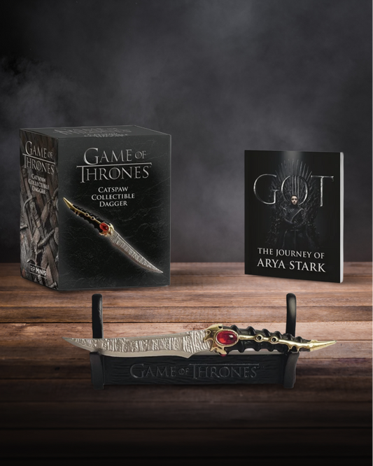 Game of Thrones collectible dagger displayed on a black stand, with its branded box and a book titled 'The Journey of Arya Stark' arranged on a dark wooden surface against a smoky gray background.
