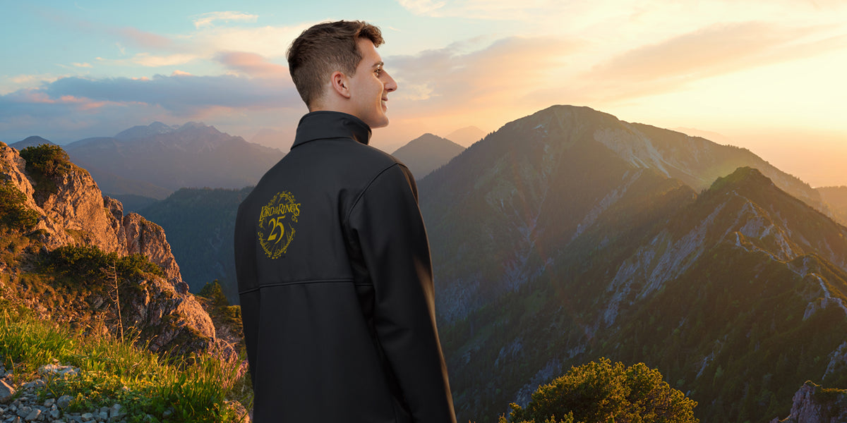 A man stands on a mountain ridge at sunset, wearing a black jacket with a gold crest logo on the back. The background features dramatic mountain peaks under a warm golden sky.